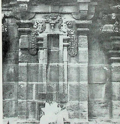 The side wall of a stone temple with a central niche framed by pilasters and curling makara-arch ornament, under a cornice of miniature roofs; fallen material at the foot.