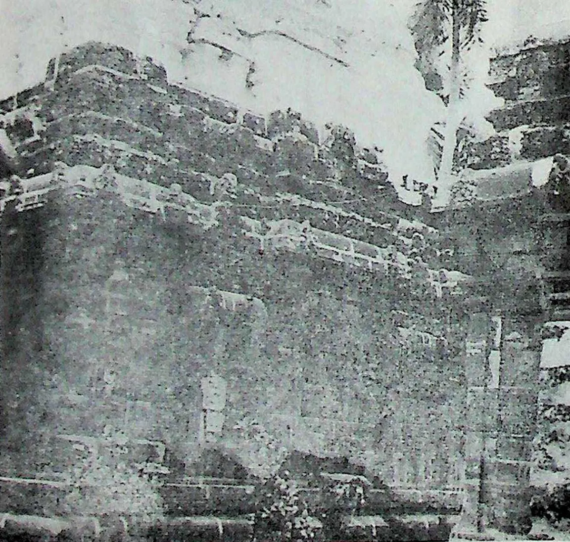 The long side wall of a weathered stone temple, its cornice and upper storeys broken and overgrown, seen at an angle against a pale sky.