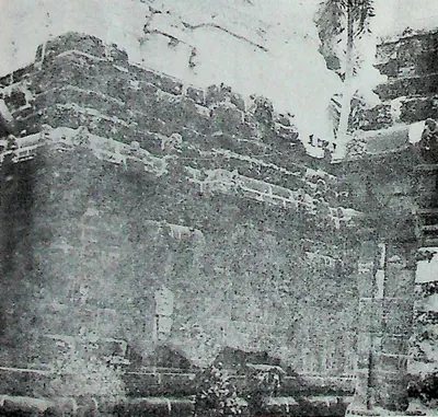The long side wall of a weathered stone temple, its cornice and upper storeys broken and overgrown, seen at an angle against a pale sky.