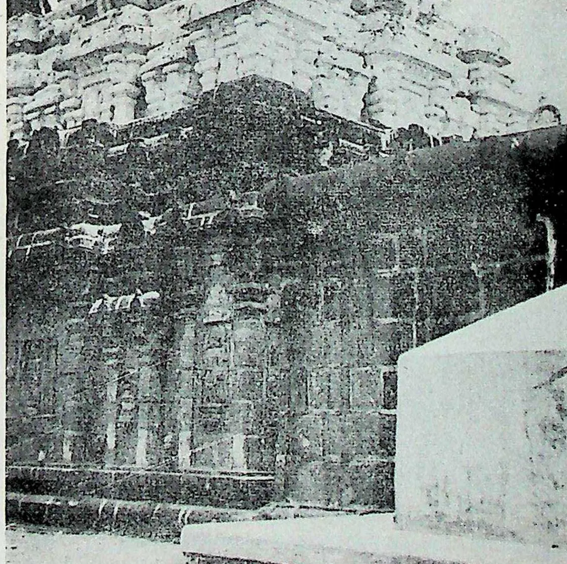 The dark side wall of a stone temple with pilasters and niches, the carved tower above, and a whitewashed later wall in the right foreground.