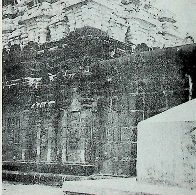 The dark side wall of a stone temple with pilasters and niches, the carved tower above, and a whitewashed later wall in the right foreground.