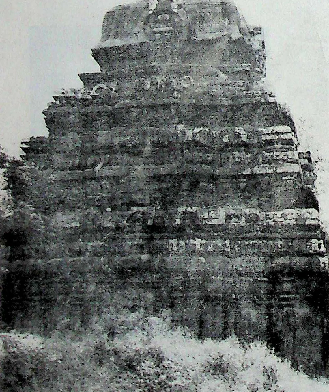 The weathered stepped tower and upper walls of a stone temple, seen close and slightly from below, with grass and scrub in front.