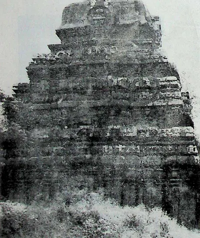 The weathered stepped tower and upper walls of a stone temple, seen close and slightly from below, with grass and scrub in front.