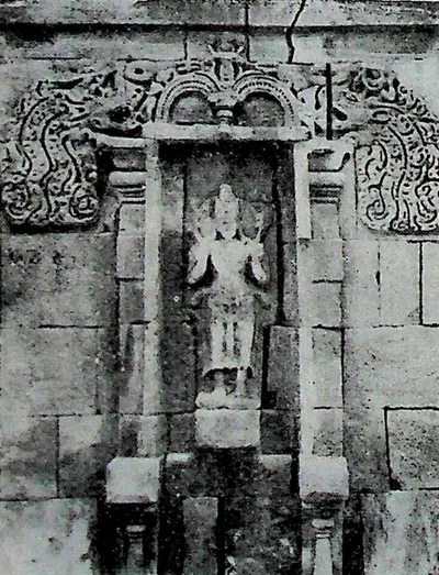 A deep wall niche with an ornate arched surround of scrolls and makara heads, containing a small standing figure identified as Surya.