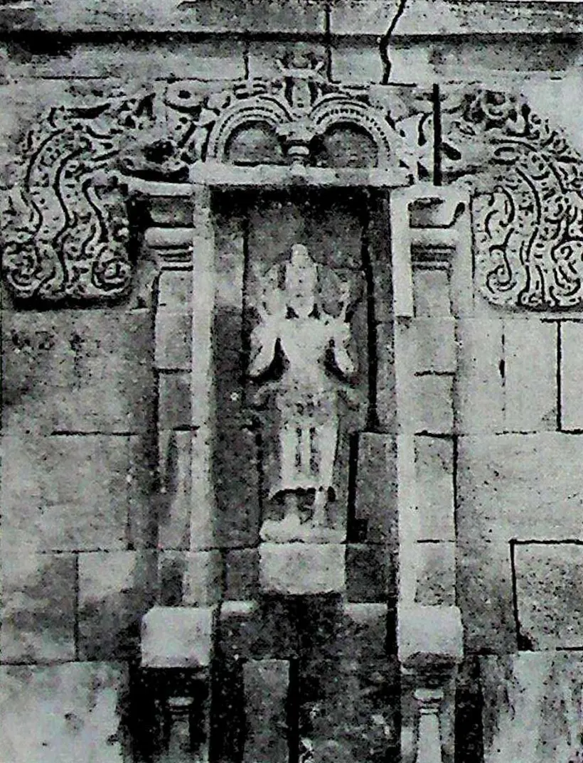 A deep wall niche with an ornate arched surround of scrolls and makara heads, containing a small standing figure identified as Surya.