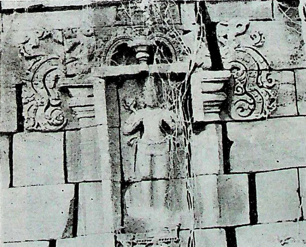A wall niche in jointed masonry with scroll ornament at the top corners, containing a standing figure identified as Surya; cracks and stains across the blocks.