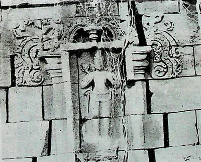 A wall niche in jointed masonry with scroll ornament at the top corners, containing a standing figure identified as Surya; cracks and stains across the blocks.
