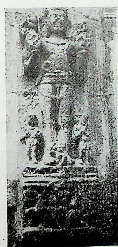 A narrow upright relief of a standing male figure, identified as Surya, with two small attendant figures at his feet, above a dark plinth.