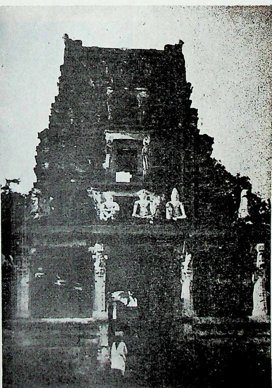 A dark, tall gateway tower with small figures on its upper storey and a doorway at the base, where a person in white stands.