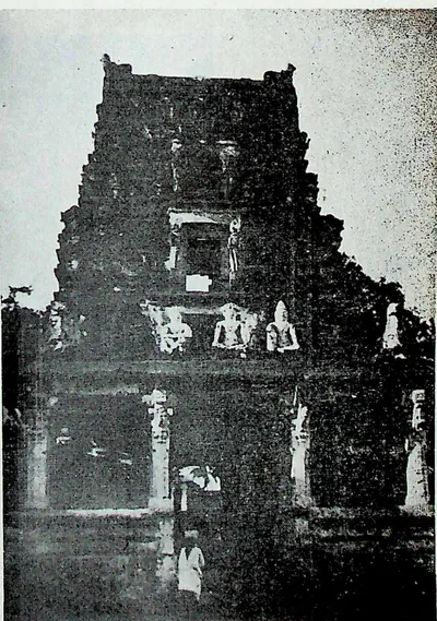A dark, tall gateway tower with small figures on its upper storey and a doorway at the base, where a person in white stands.