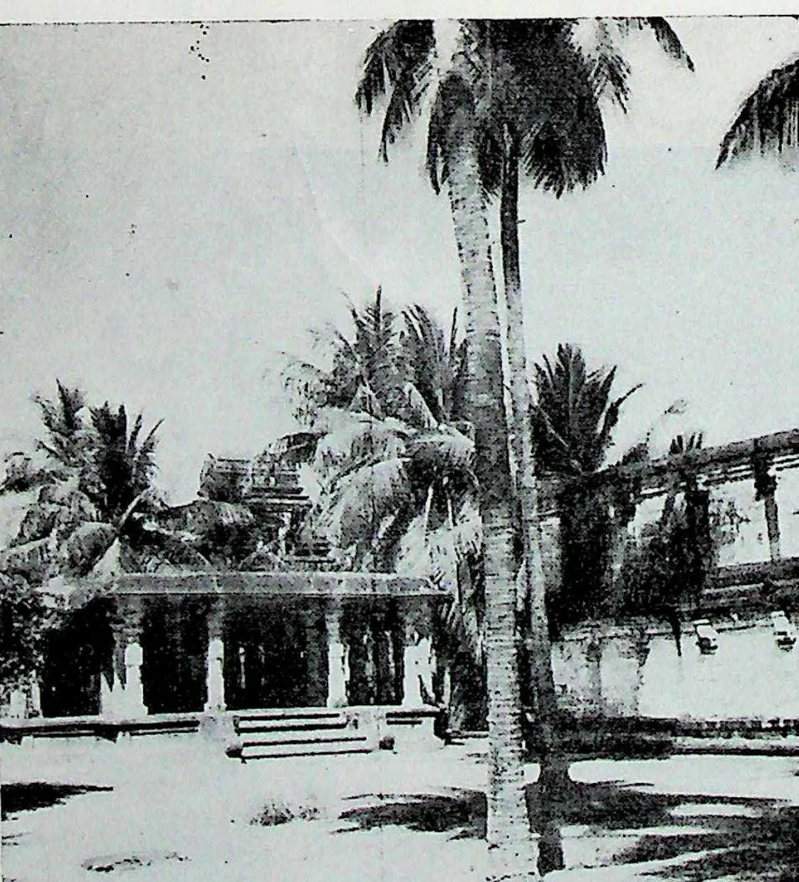 A low pillared hall in the corner of the temple enclosure, seen across a courtyard with tall coconut palms.