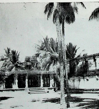 A low pillared hall in the corner of the temple enclosure, seen across a courtyard with tall coconut palms.