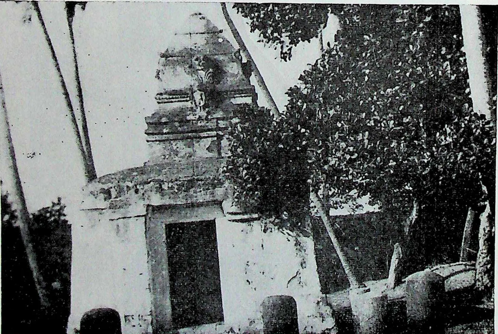 A small whitewashed shrine with a dark doorway and a low tower, shaded by trees and palm trunks.