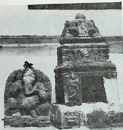 A miniature stone shrine with a domed top beside a seated Ganesha image, in front of a long wall.