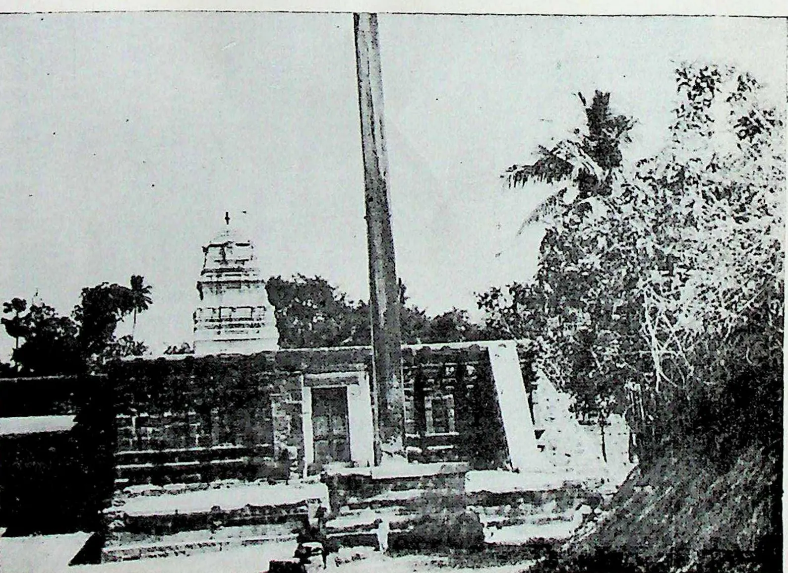 The western entrance of the outer enclosure at Chalukya Bhimavaram (Samalkot): a low gateway and wall with a tall flagstaff, a small tower behind, and trees.