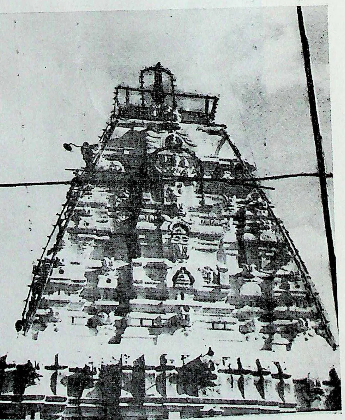 A tall pyramidal tower of many tiers covered with small figures and roof mouldings, with wires crossing in front.