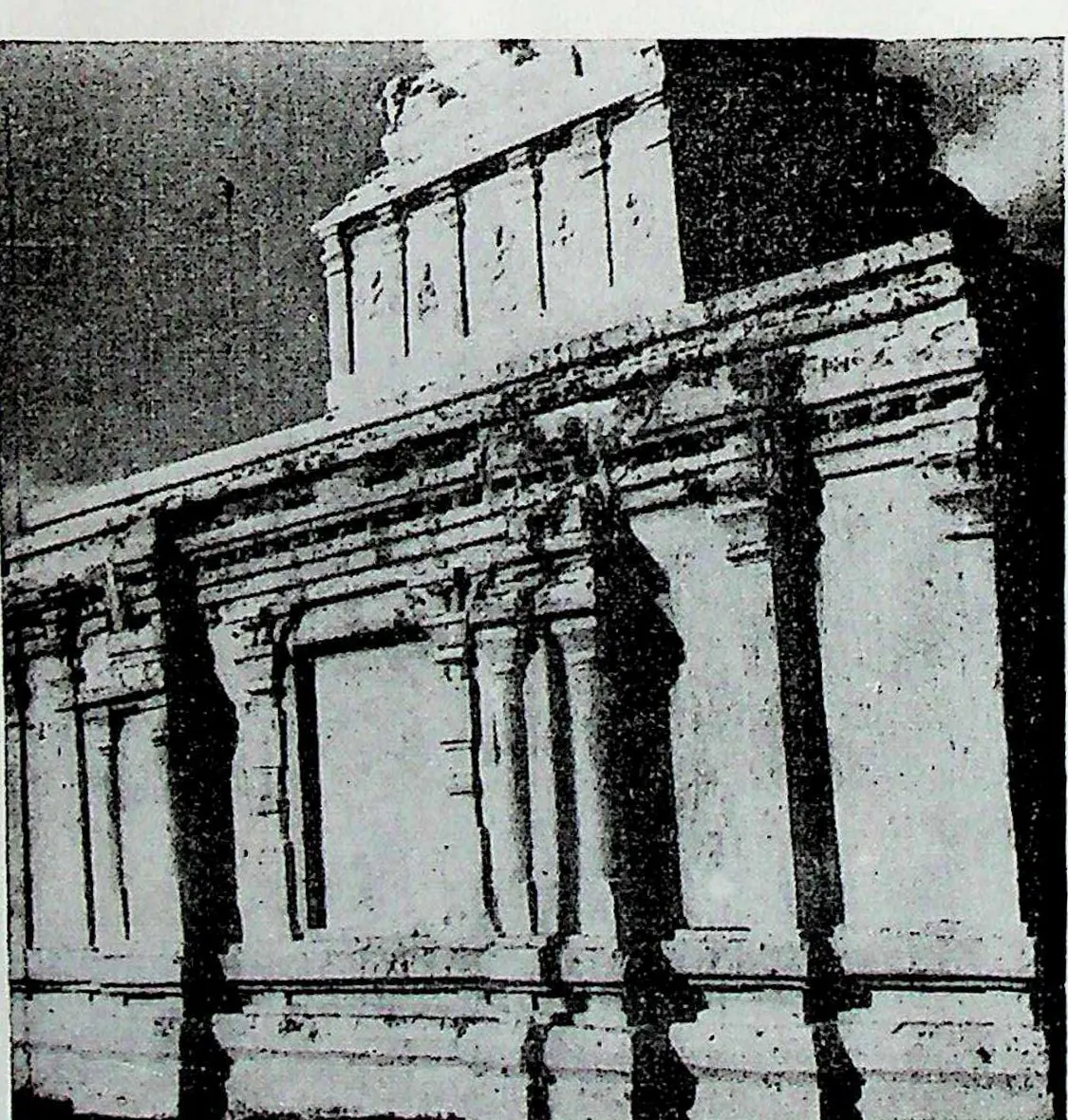A whitewashed temple wall of pilasters and niches under a heavy cornice, with a small tower above, seen in strong light and shadow.