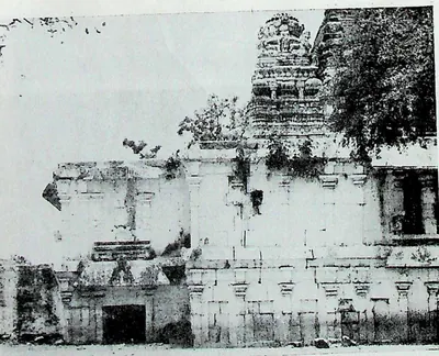 The north side of a temple: a long low wall with niches and a tower rising behind, overgrown in places.
