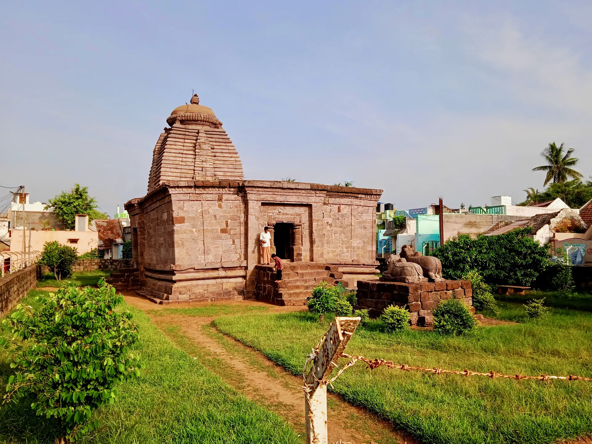 Colour photograph of a small stone temple with a curved tower over the shrine and a flat-roofed hall in front, standing in a garden of grass and shrubs.