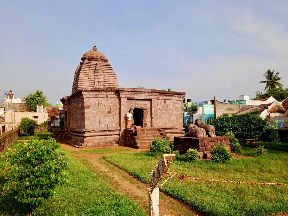 Colour photograph of a small stone temple with a curved tower over the shrine and a flat-roofed hall in front, standing in a garden of grass and shrubs.