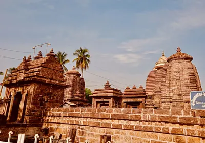 Colour photograph of a paved temple courtyard of red-brown stone: small shrines with curved, ribbed towers and stepped roofs stand along a low enclosure wall under a blue sky with palm trees.
