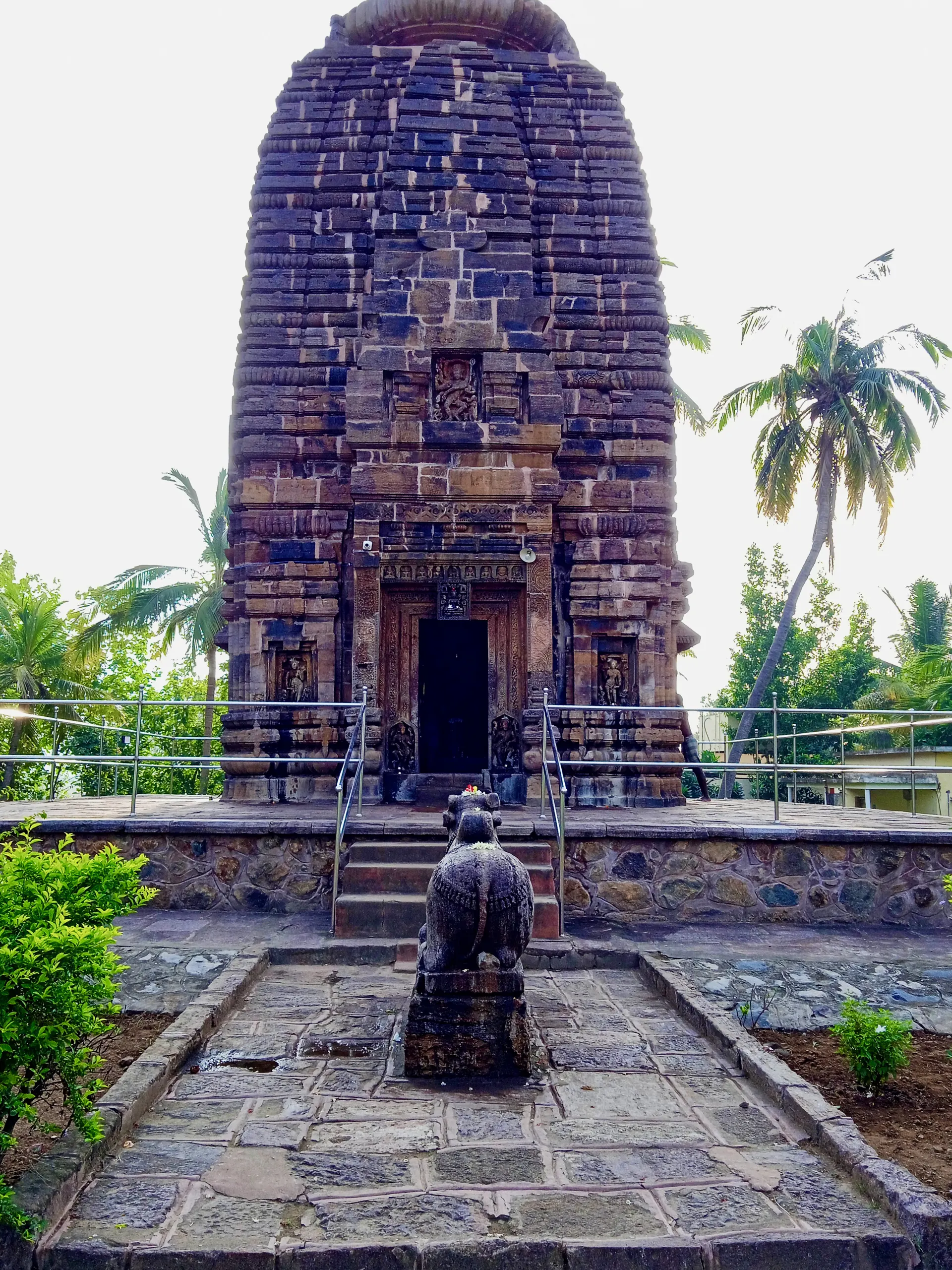 Colour photograph of a tall stone temple tower of many thin horizontal courses rising over a doorway, with a seated stone bull on a platform in front and palm trees beside it.