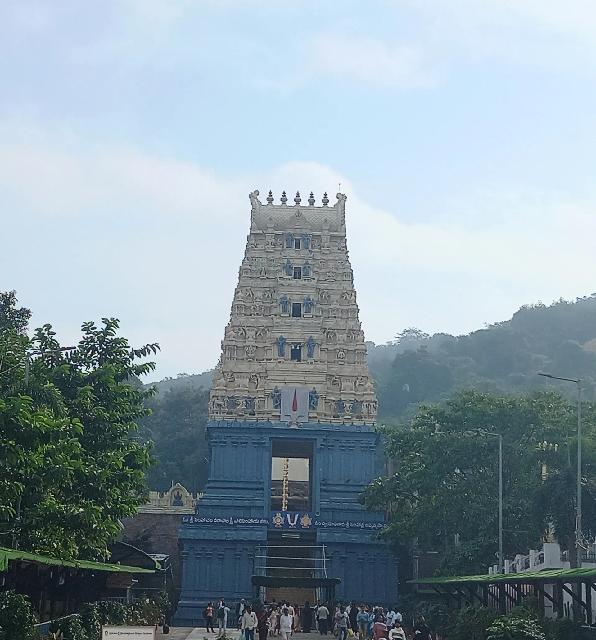 Colour photograph of a tall, tiered temple gateway tower painted in pale colours, seen from below over the heads of visitors, with forested hills behind.