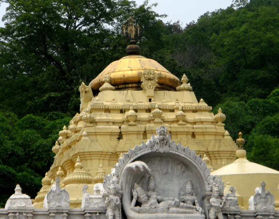 Colour photograph of the domed tower over the shrine of the Simhachalam temple, painted gold and white, with a large stucco figure in an arched niche on its face and wooded hills behind.