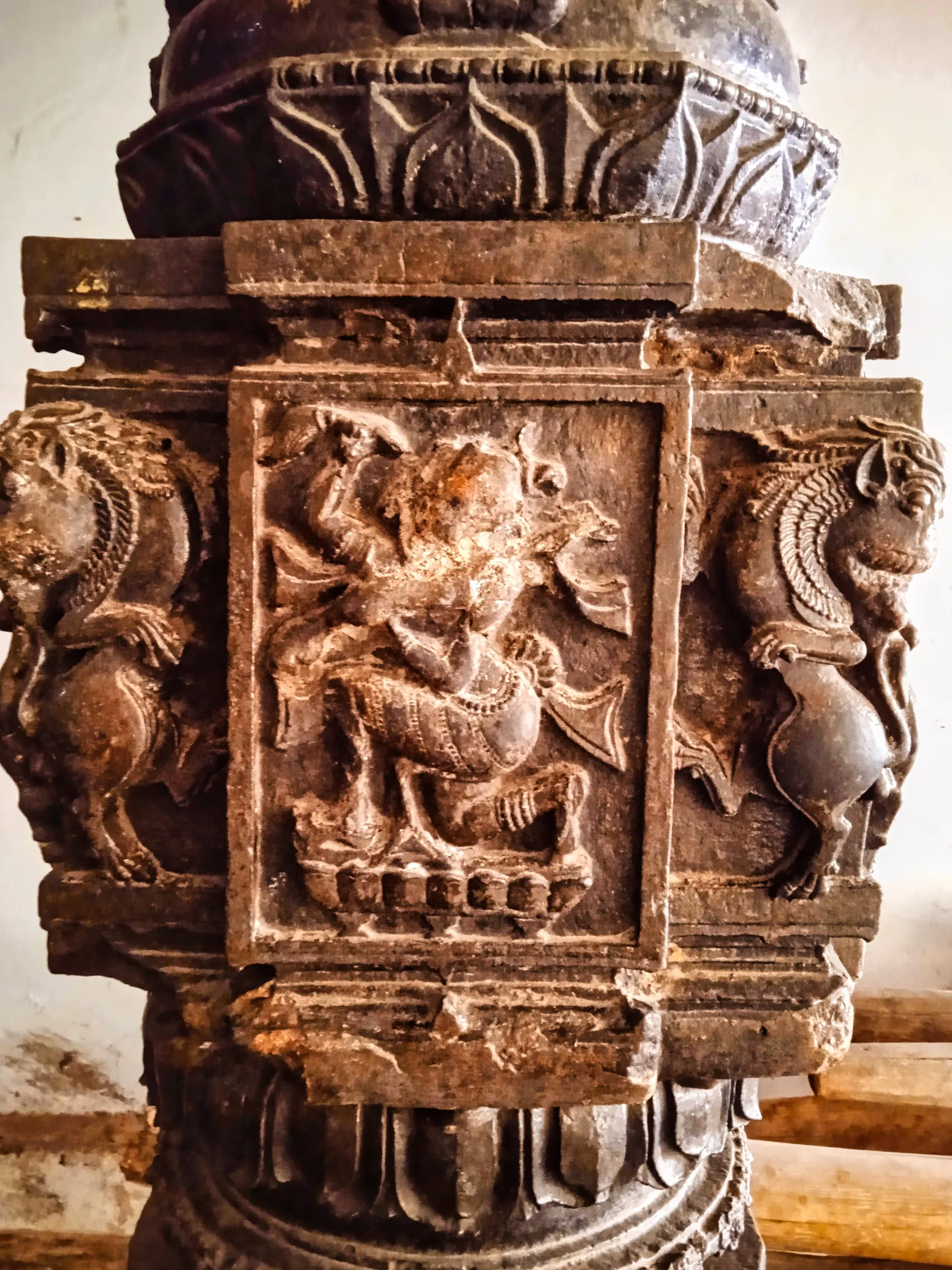 Colour close-up photograph of a dark stone pillar block carved with a figure in a square panel, flanked by rearing animal brackets.