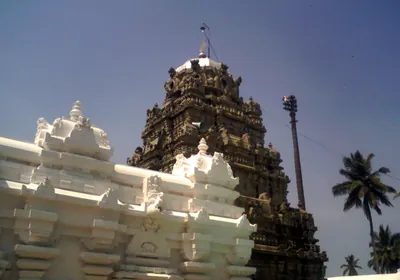Colour photograph of a temple seen from outside its whitewashed enclosure: a dark stone tower with sculpture rises behind a white parapet against a blue sky.