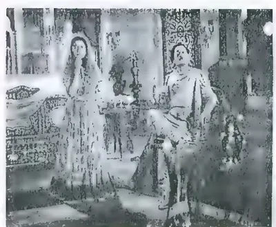 Black-and-white film still of a woman in a sari standing with a hand at her face, and a man seated on a carved chair in a decorated room.