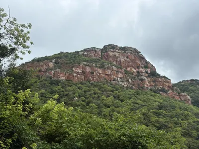 A steep wooded hill rising to bare reddish cliffs under a cloudy sky.