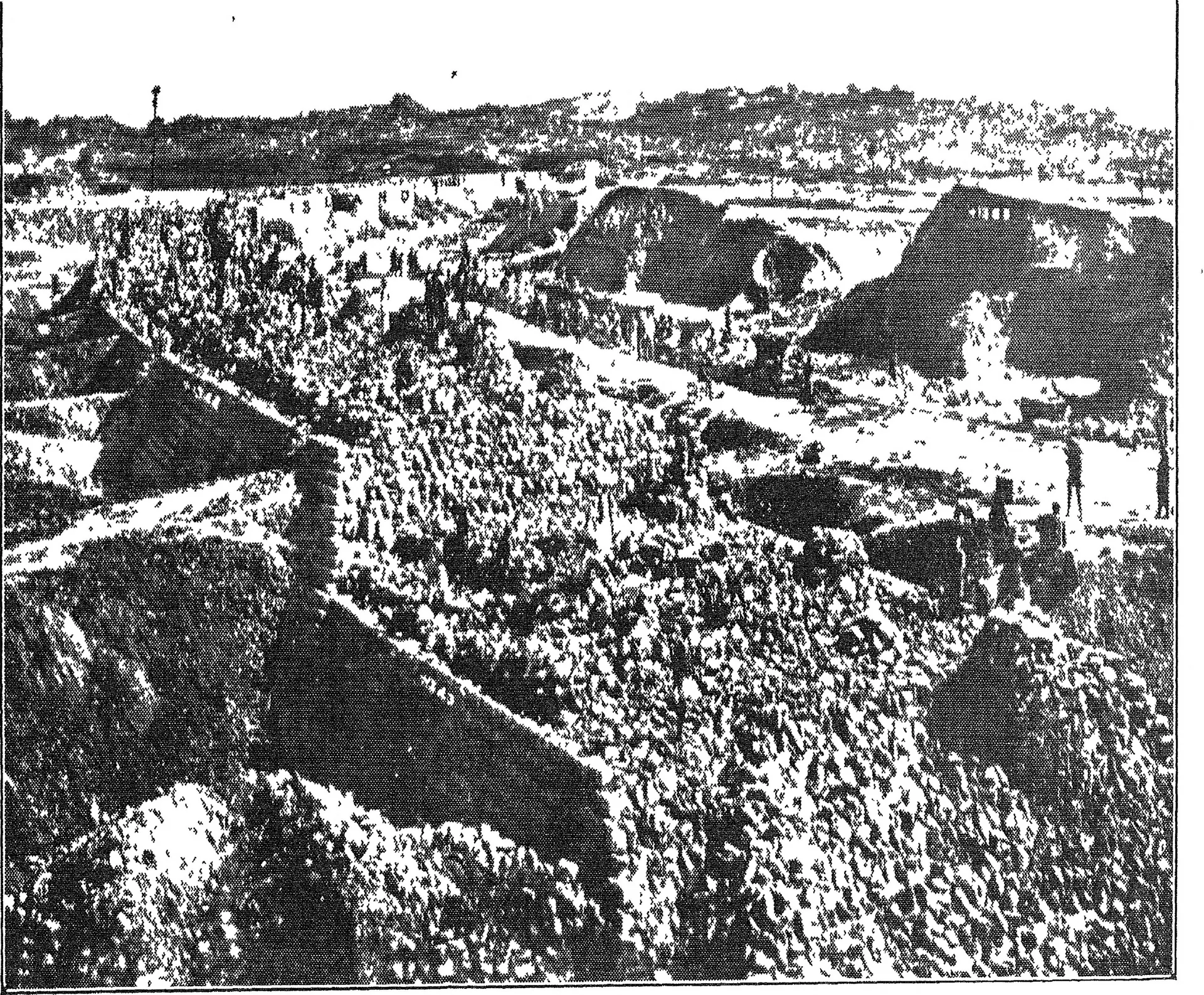 Old halftone photograph of a long masonry dam across a rocky river valley, with buildings and small figures on top and hills behind.