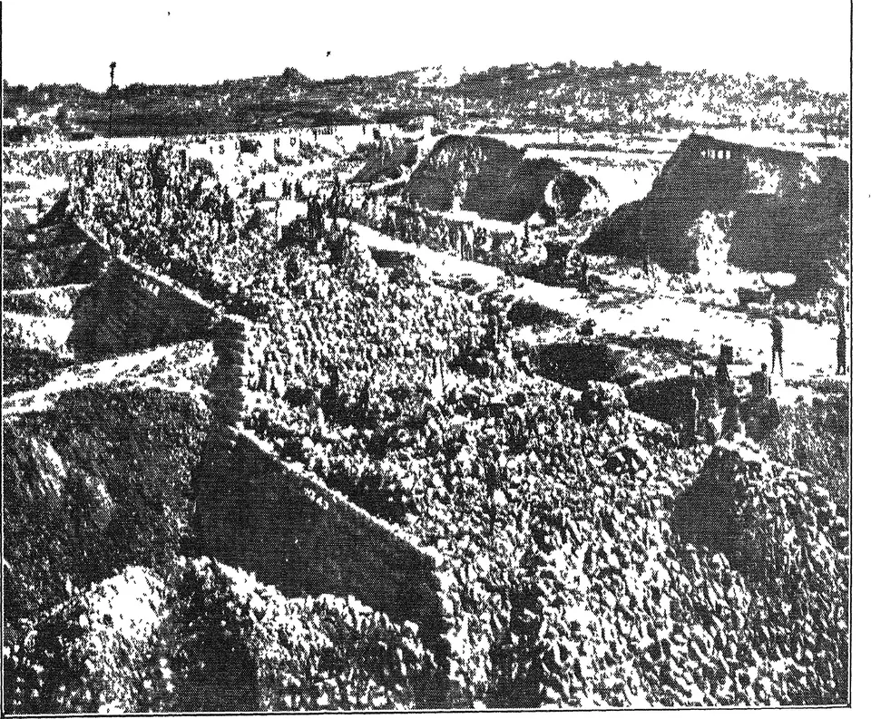 Old halftone photograph of a long masonry dam across a rocky river valley, with buildings and small figures on top and hills behind.