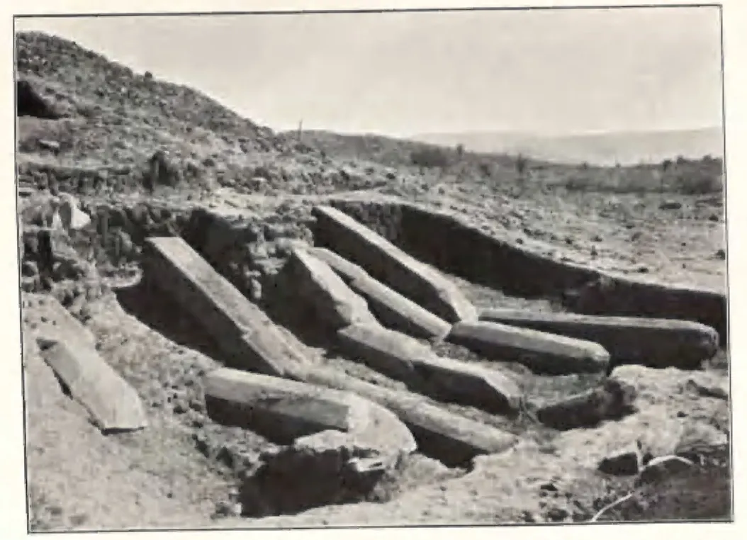 Long fallen octagonal stone pillars lying in a trench beside the stupa, with bare hills behind.