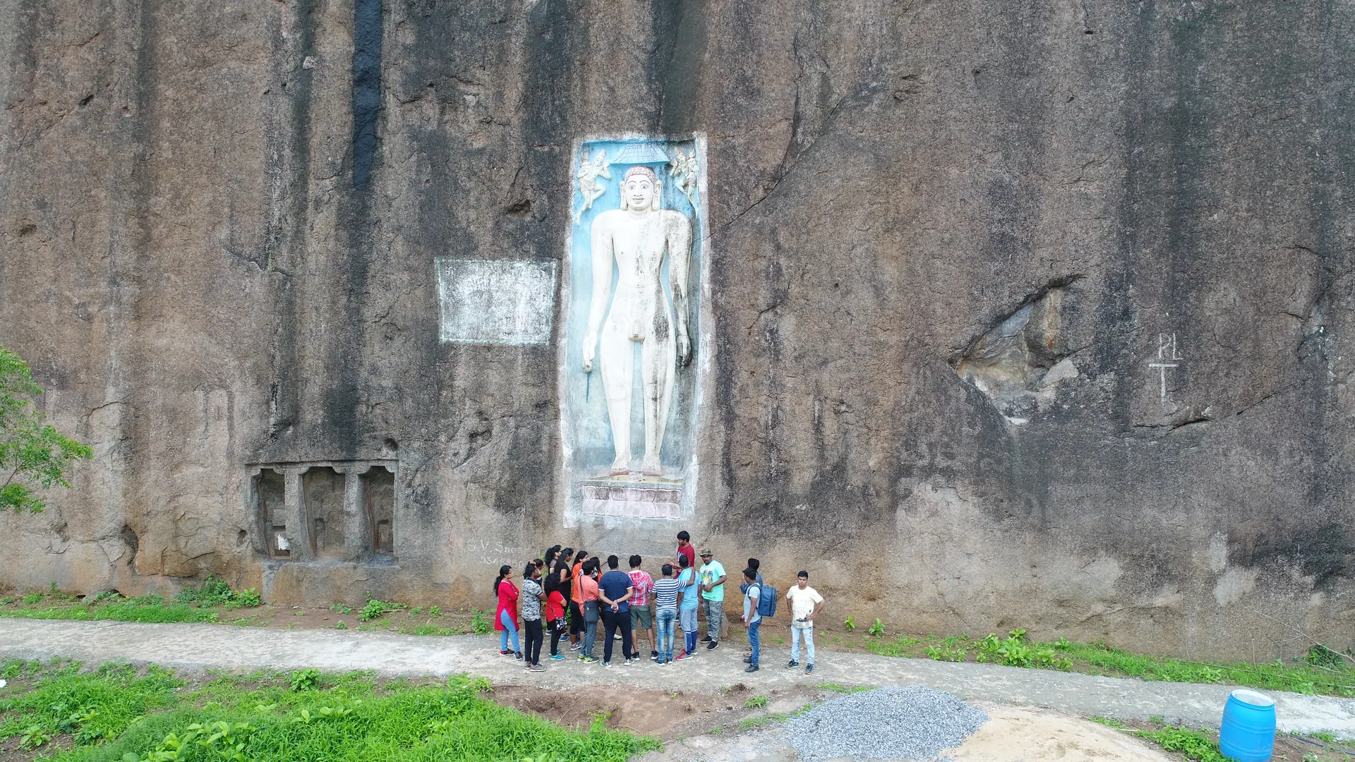 A sheer dark granite rock face with a tall standing Jain figure carved in a sunken panel and painted white on blue, a group of visitors on the path at its foot.