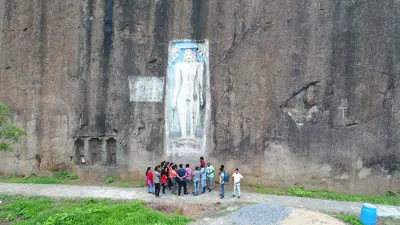 A sheer dark granite rock face with a tall standing Jain figure carved in a sunken panel and painted white on blue, a group of visitors on the path at its foot.