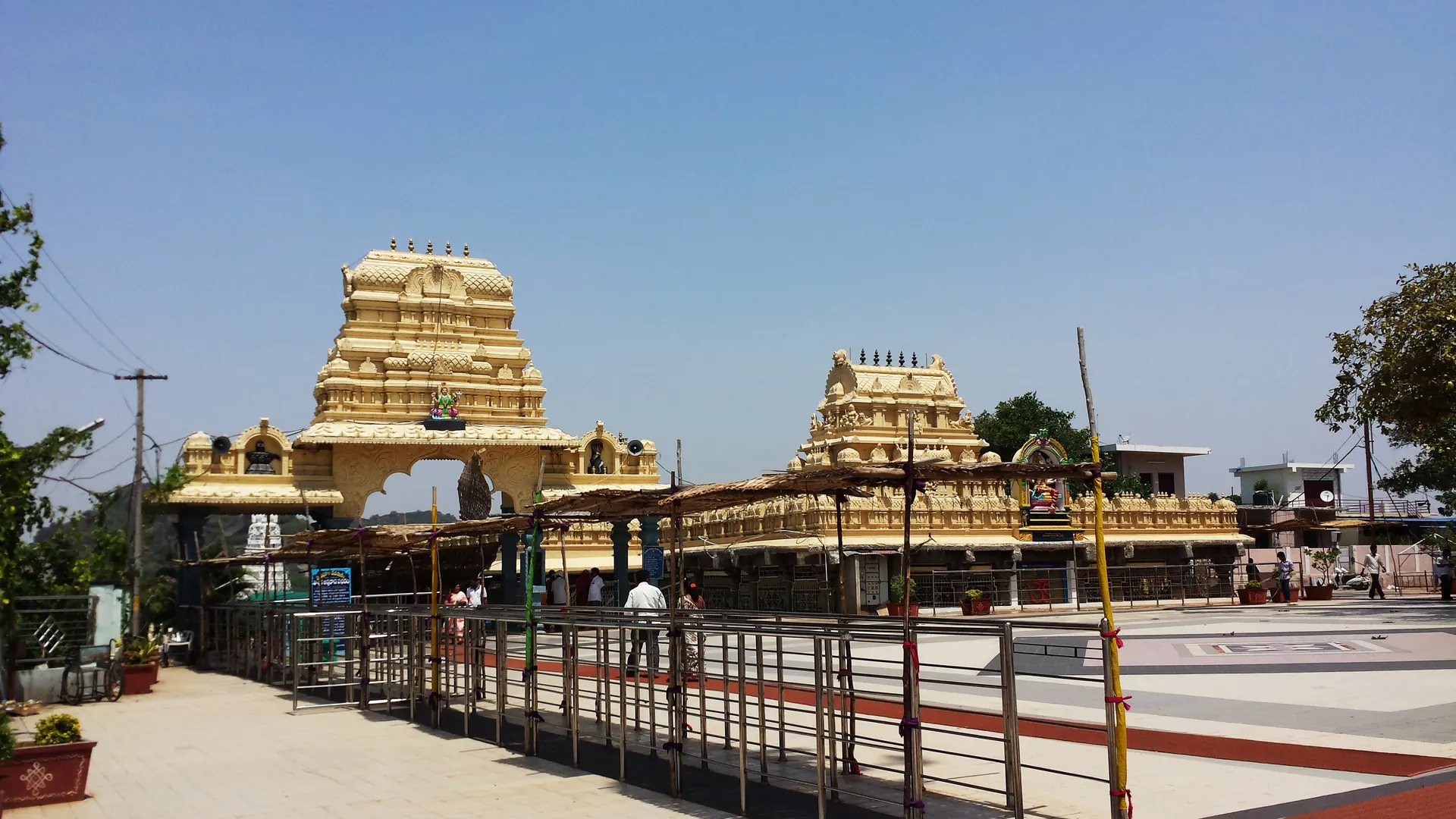 Two cream-and-gold tiered gateway towers rising above a long, low pillared hall, with a paved forecourt and metal queue railings in front.