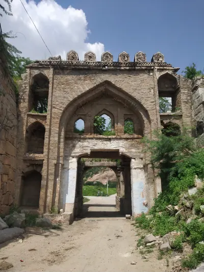 A tall stone gateway with a large pointed arch over a smaller square doorway, three small arched openings inside the arch and a dirt road passing through.
