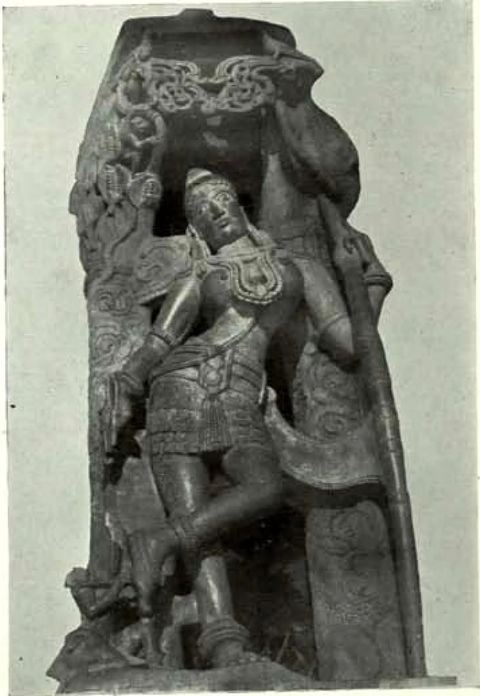 A stone bracket carved with a crowned, jewelled figure in a pleated skirt standing cross-legged beside a long bow, under a carved creeper; shown cut out against a plain background.
