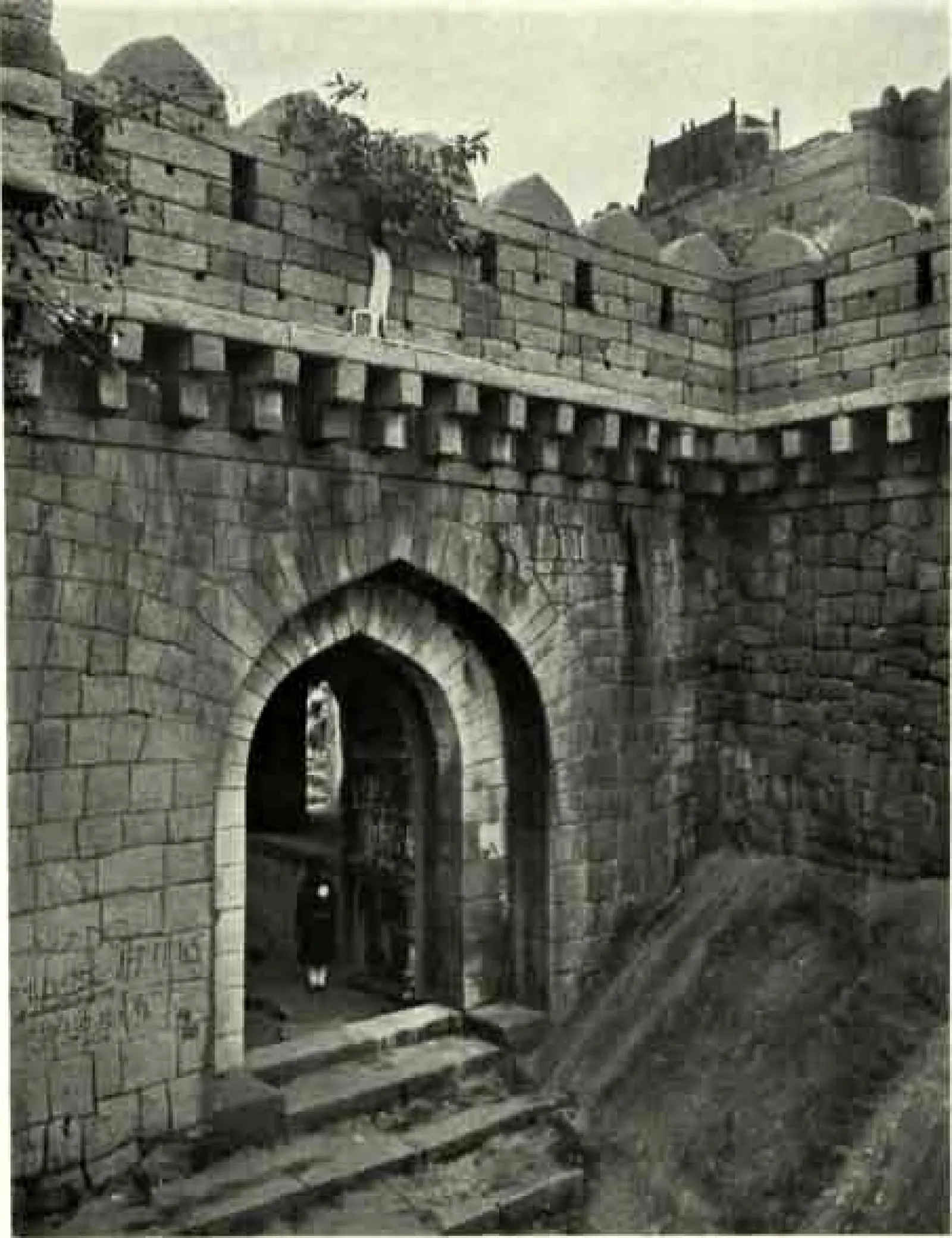 A pointed-arch gateway in a high battlemented wall of dressed stone, reached by stone steps, with a person standing in the passage.