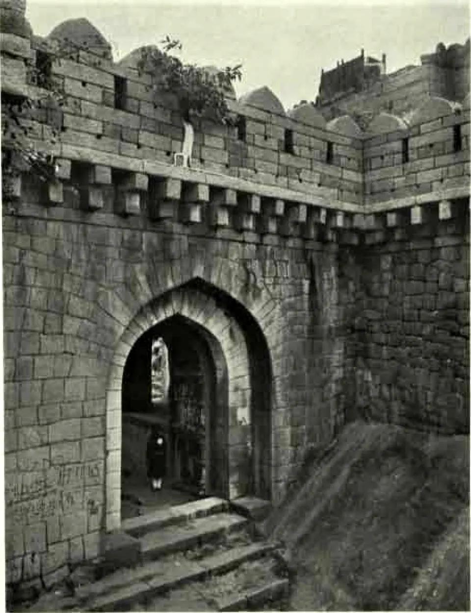 A pointed-arch gateway in a high battlemented wall of dressed stone, reached by stone steps, with a person standing in the passage.
