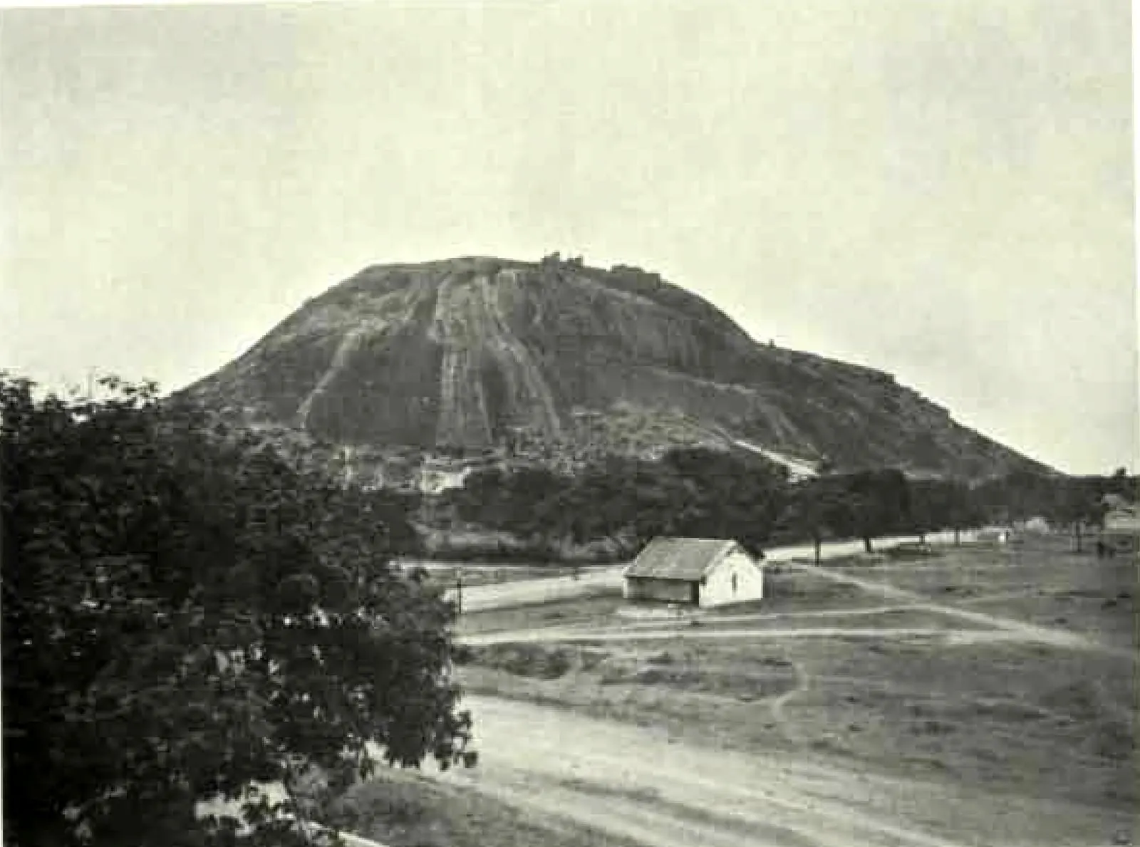 A great bare granite hill rising above trees and an open plain, with a road and a small building in the foreground.