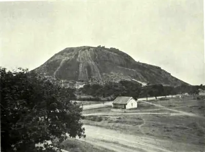 A great bare granite hill rising above trees and an open plain, with a road and a small building in the foreground.
