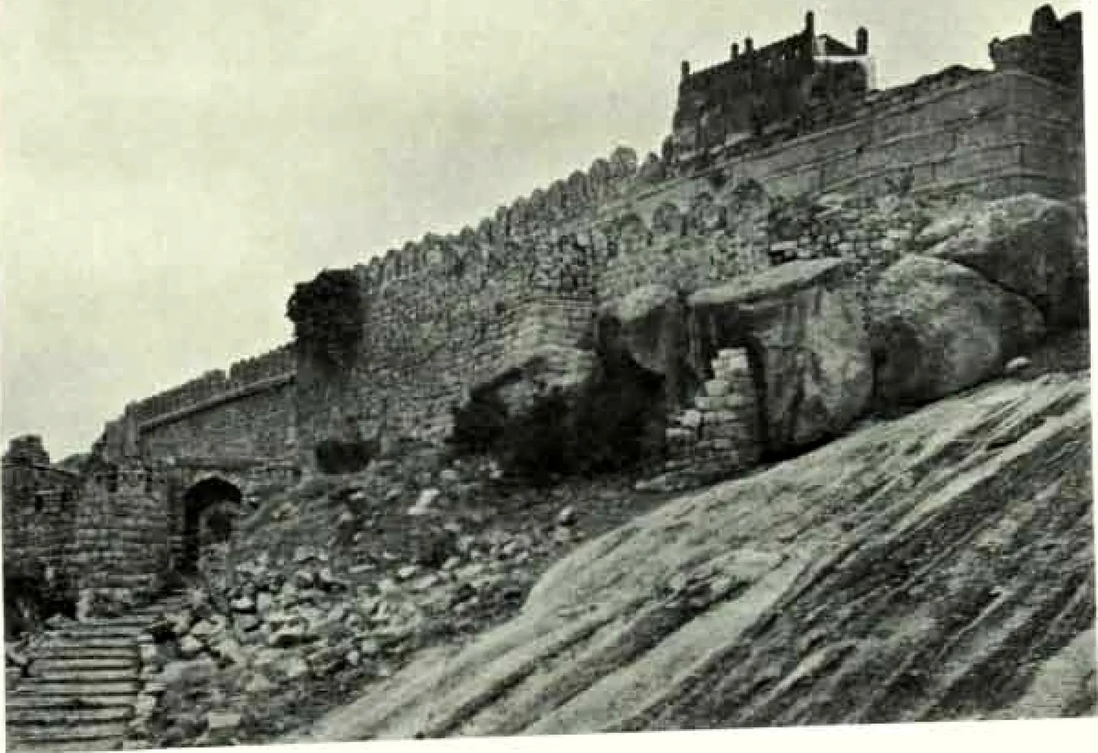 A battlemented stone wall climbing a sloping sheet of granite, with steps and a small gateway at the lower left.