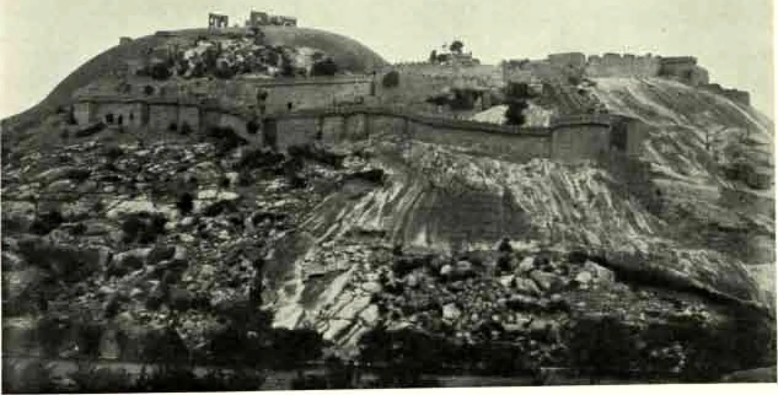 Ruined walls and buildings along the top of the granite rock, seen from below across rough, boulder-strewn ground.