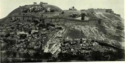 Ruined walls and buildings along the top of the granite rock, seen from below across rough, boulder-strewn ground.