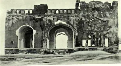 The ruined front of a building with three arched openings, a pedimented doorway at the right and a row of small openings along the top.