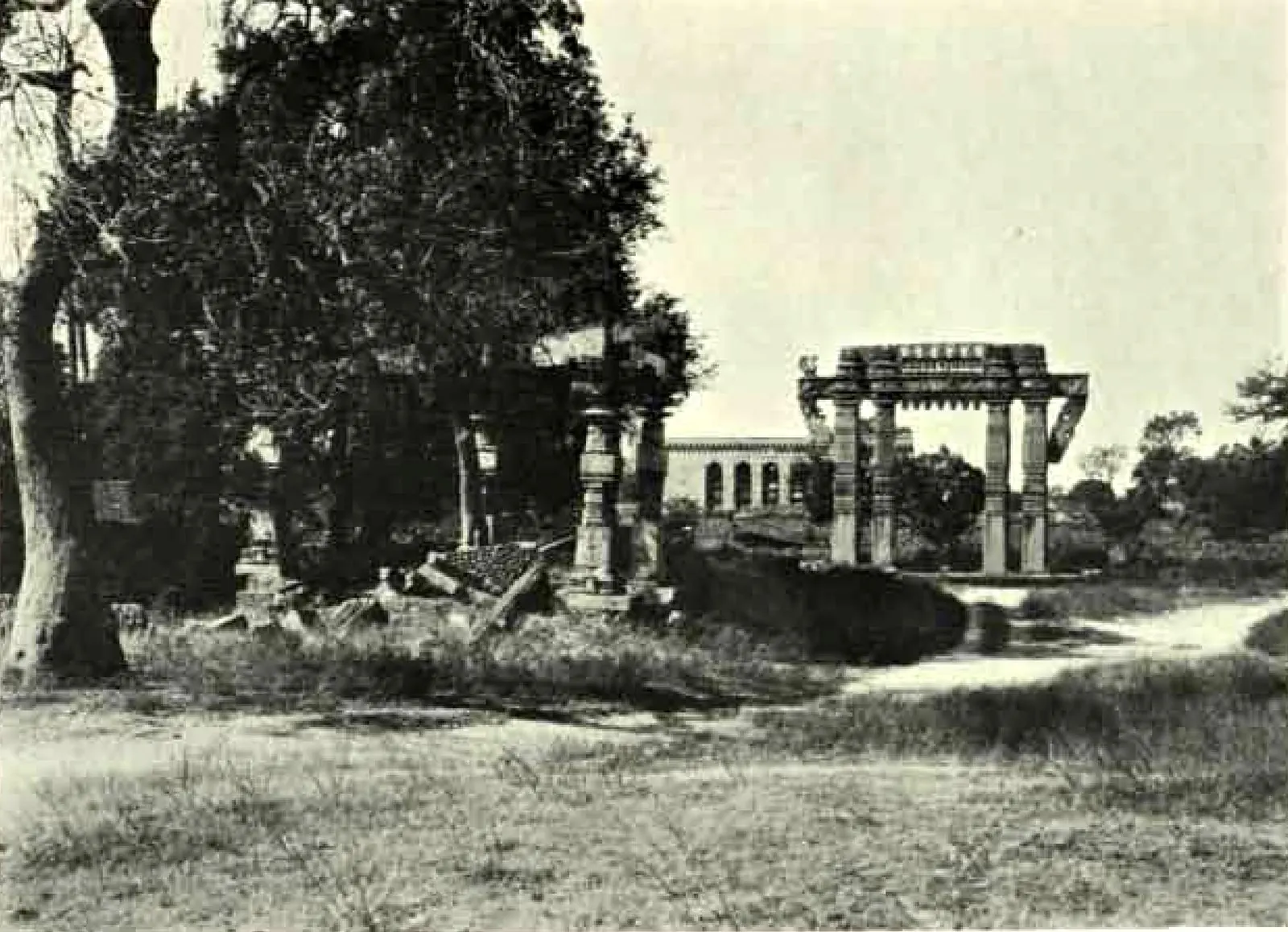 A carved stone gateway standing among grass and trees, with fallen temple stones and a pillar at the left and an arcaded building in the distance.