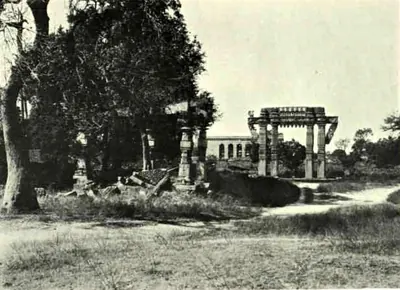 A carved stone gateway standing among grass and trees, with fallen temple stones and a pillar at the left and an arcaded building in the distance.